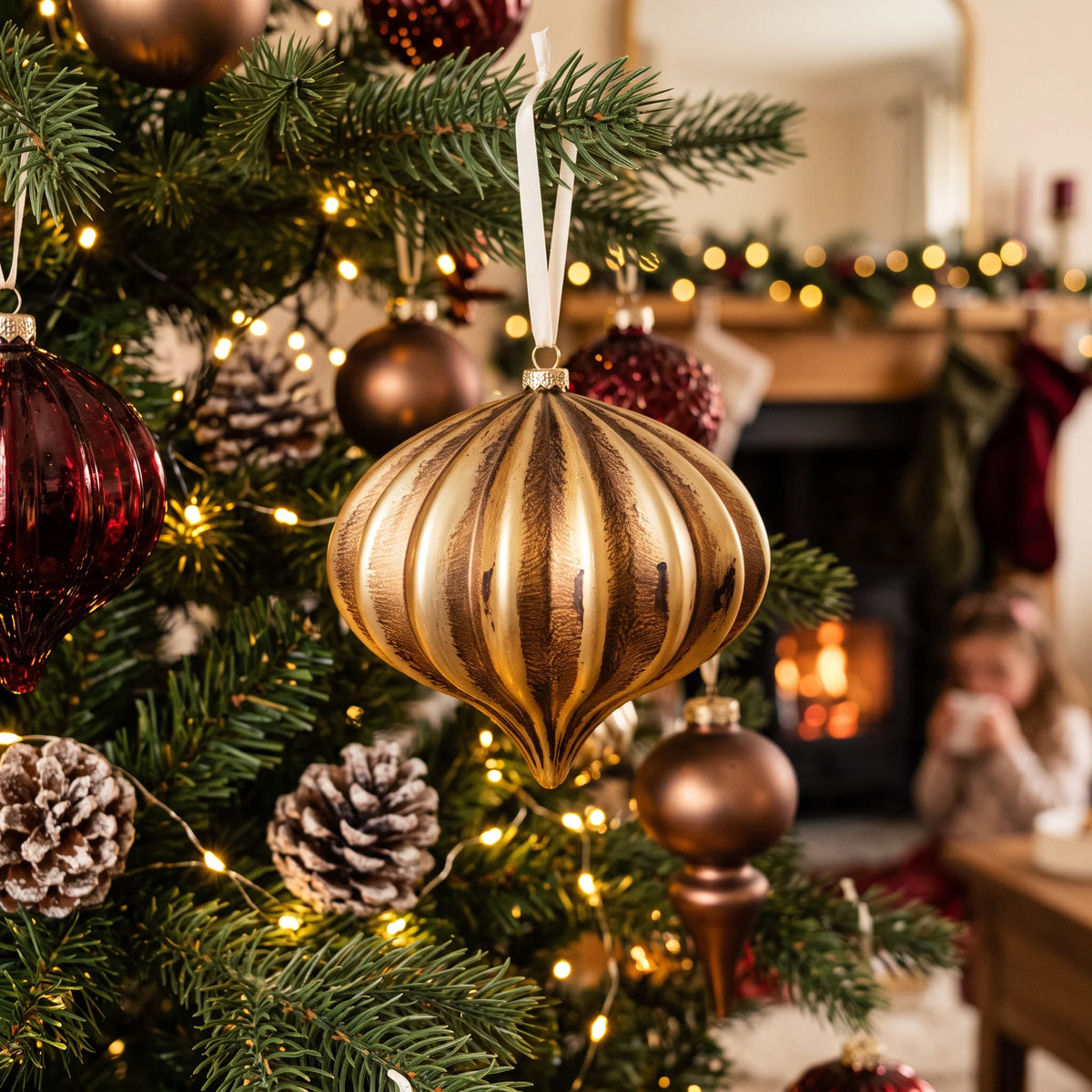Matte vintage gold onion Christmas bauble hanging on a traditional Christmas tree decorated with warm white fairy lights, pinecones and rich burgundy ornaments in a cosy festive living room.