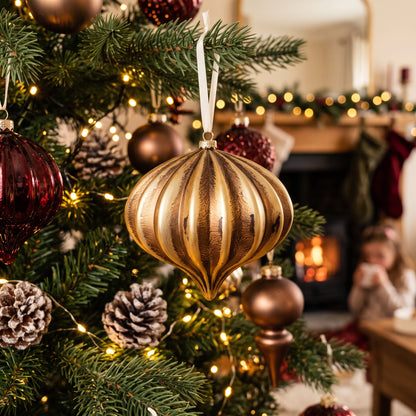 Matte vintage gold onion Christmas bauble hanging on a traditional Christmas tree decorated with warm white fairy lights, pinecones and rich burgundy ornaments in a cosy festive living room.